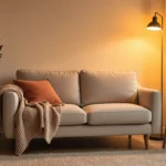 A cozy small living room corner with a compact sofa, knit throw, glowing floor lamp, and soft rug in warm neutrals.